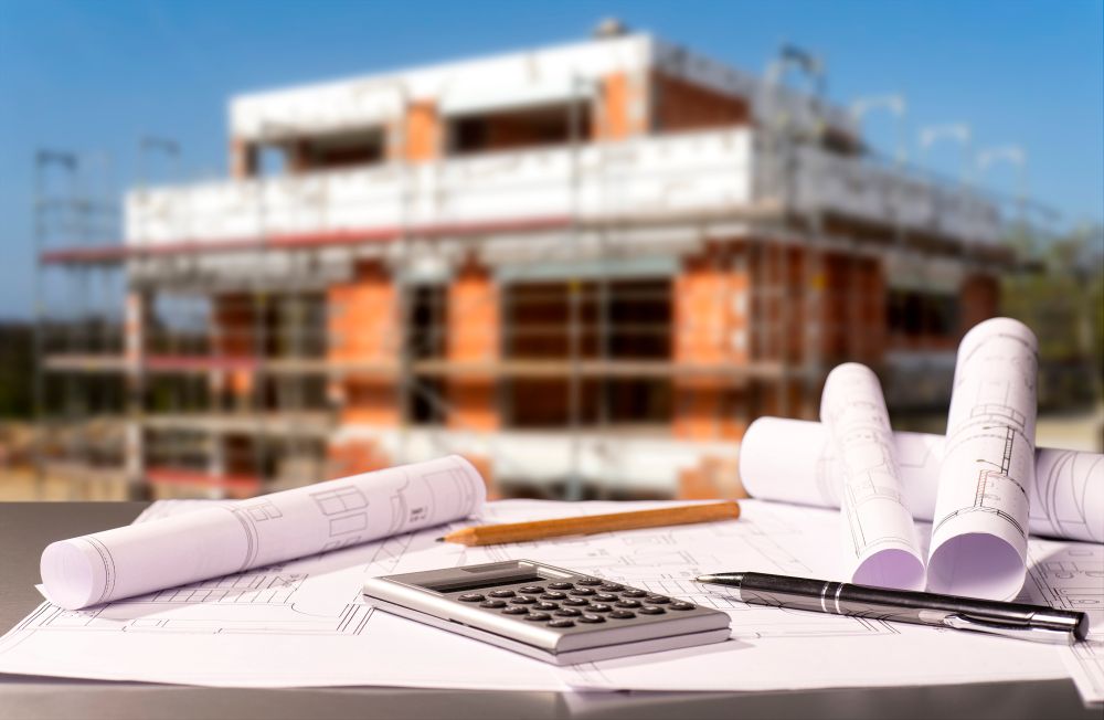bauunternehmen-in-schleswig-holstein-rohbau-bauplaene Im Vordergrund liegen die Baupläne für ein Gebäude, dessen Rohbau im Hintergrund zu sehen ist, erstellt von einem Bauunternehmen in Schleswig-Holstein.